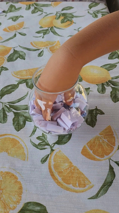 A child’s hand reaching into a glass jar filled with rolled-up summer activity strips on a citrus-themed tablecloth.