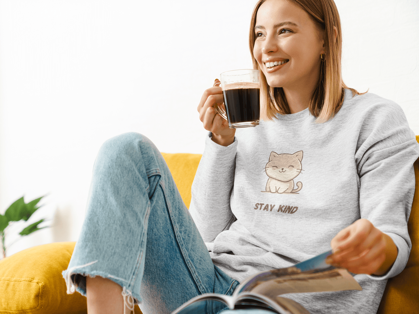 Smiling woman sitting on a yellow chair at home, wearing a gray “Stay Kind” sweatshirt with a cute cat illustration, holding a cup of coffee and reading a magazine.