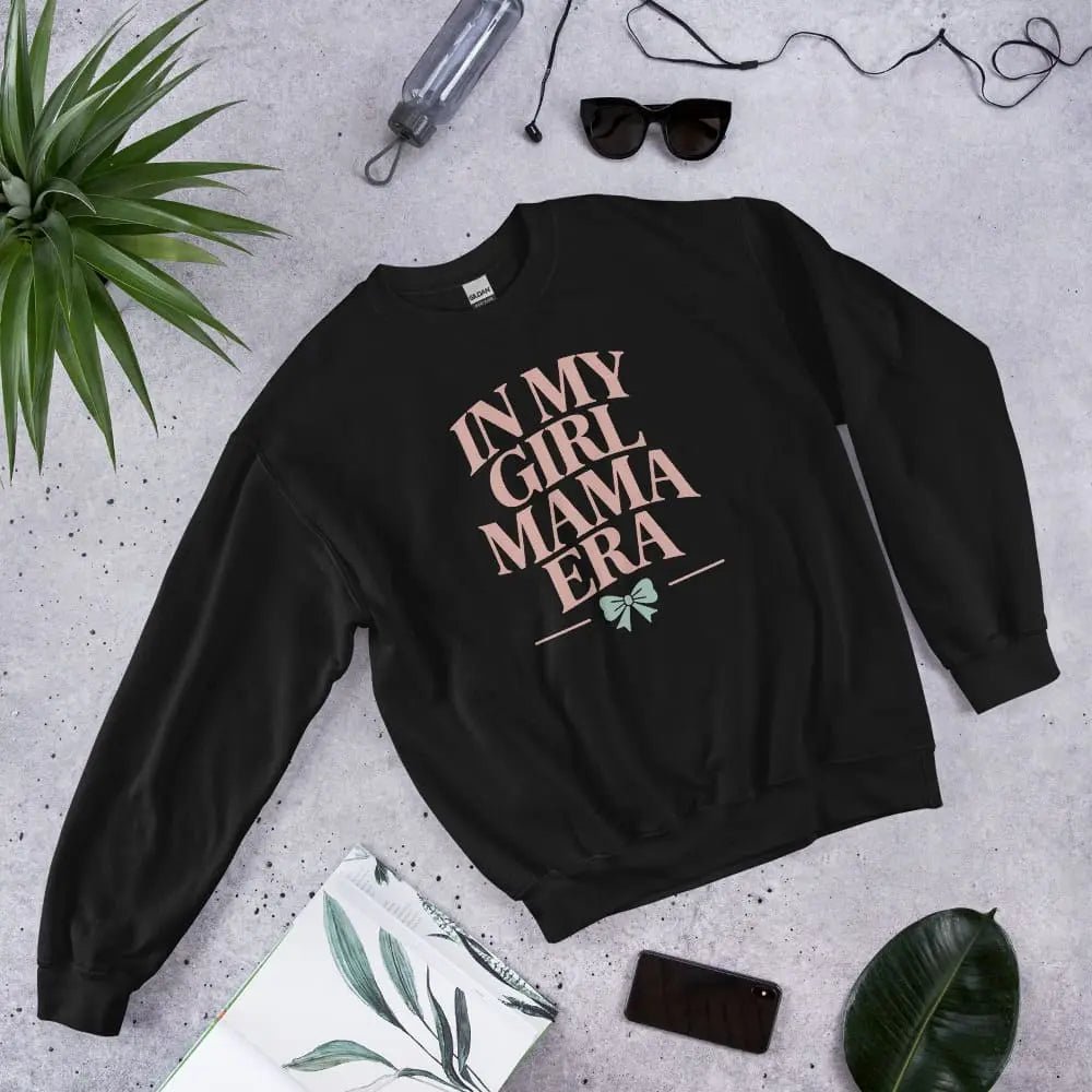 Black sweatshirt with 'In My Girl Mama Era' text on a gray surface with accessories.