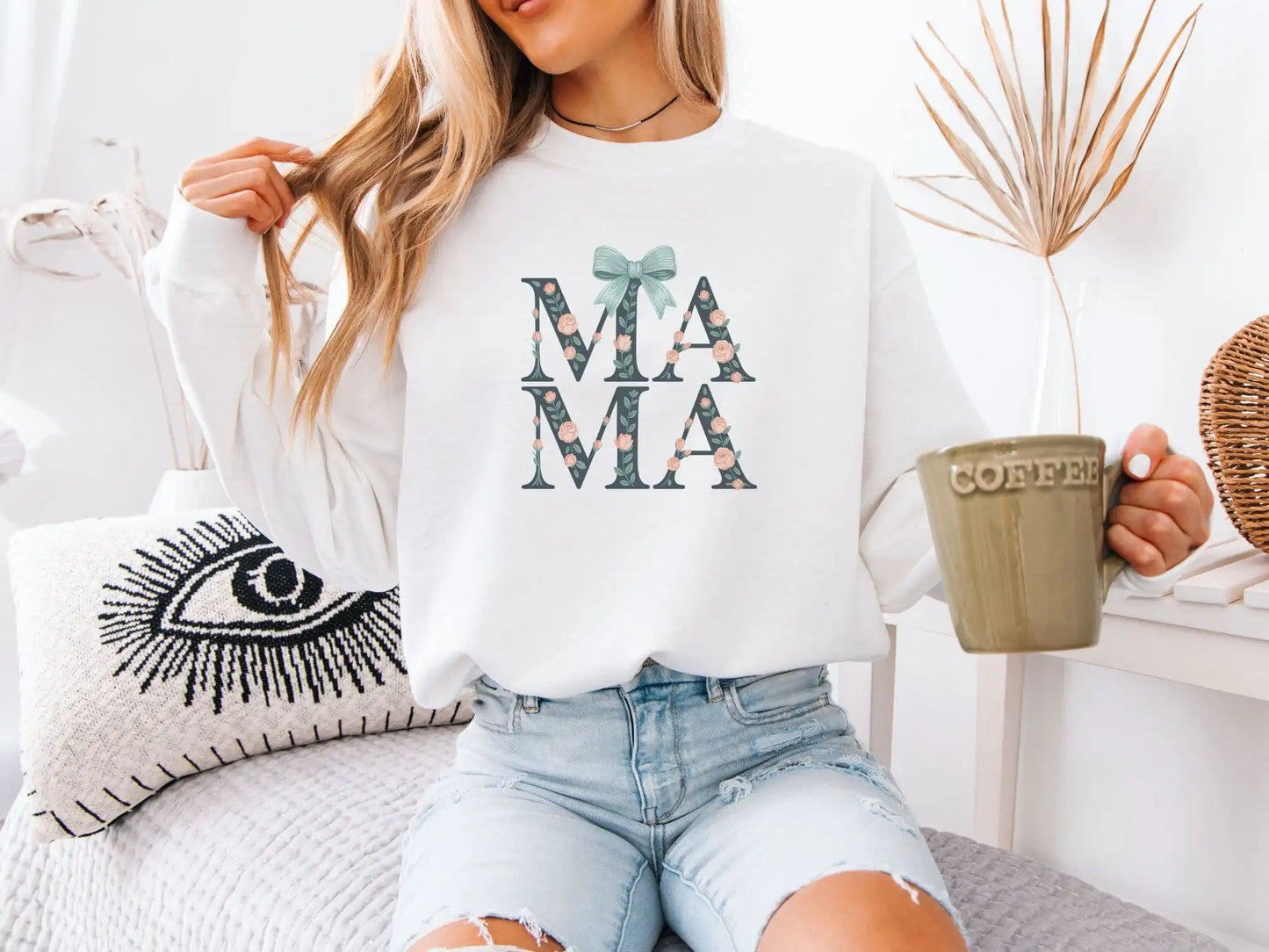 Woman wearing a white sweatshirt with 'MAMA' print, holding a mug, sitting on a couch.