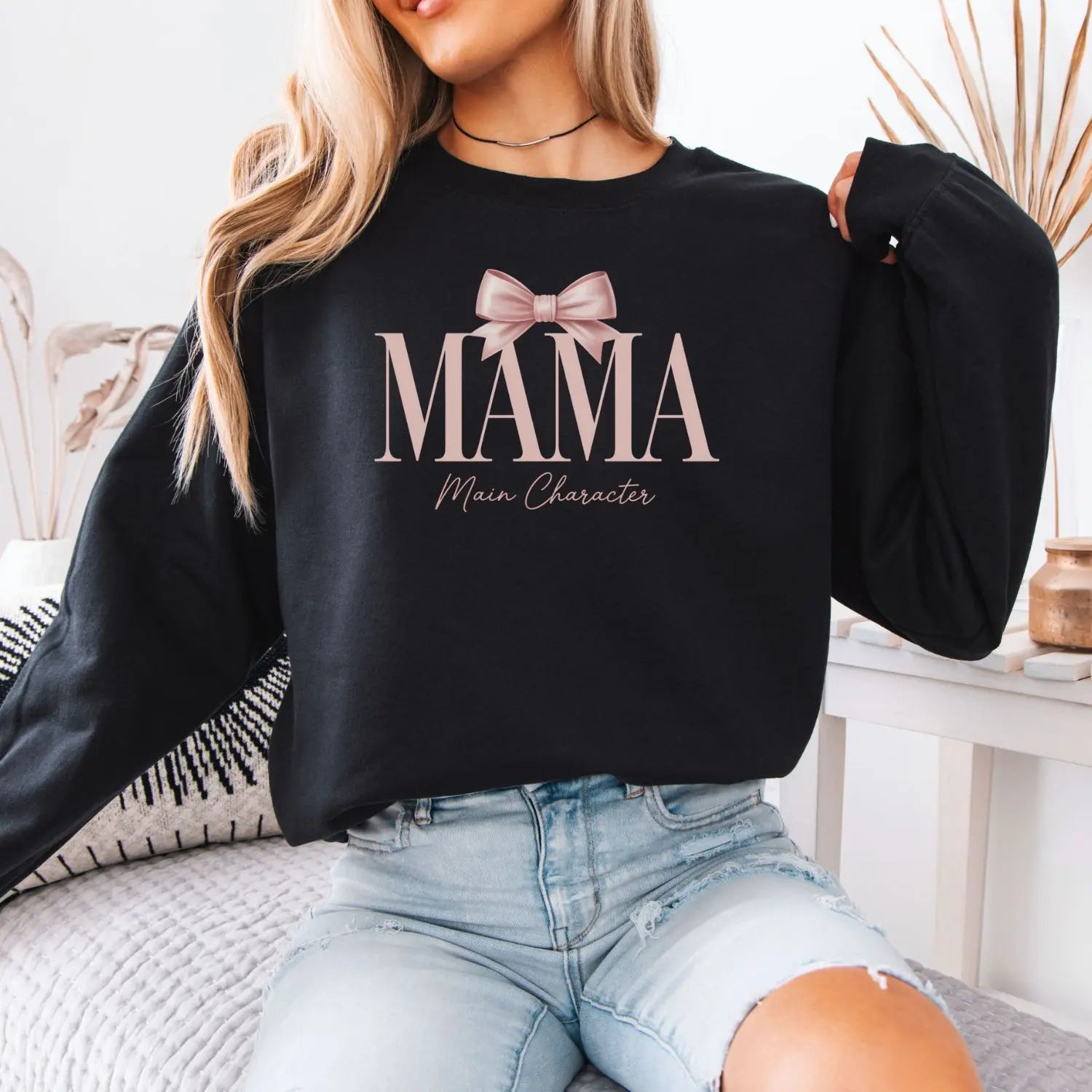 Person wearing a black sweatshirt with 'MAMA Main Character' text and a bow graphic.