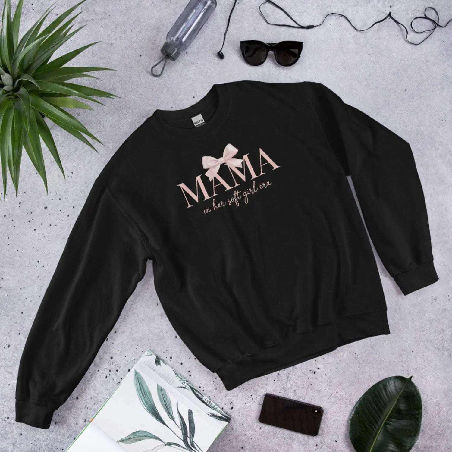Black sweatshirt with 'MAMA in her soft girl era' text on a gray surface with accessories.
