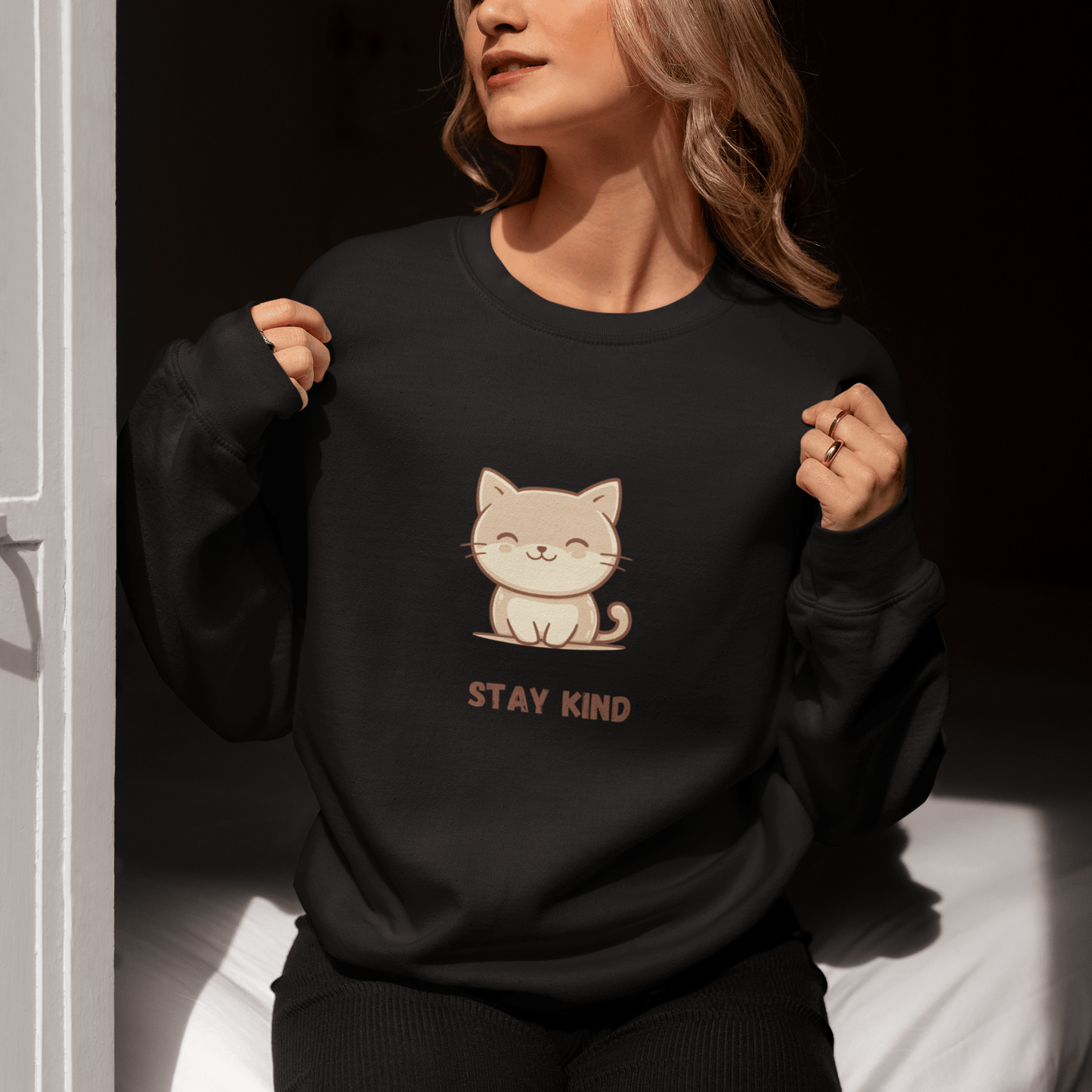 Woman sitting by a sunlit door, wearing a black sweatshirt with a cute cat illustration and the phrase “Stay Kind,” holding the collar with both hands and enjoying the sunlight.