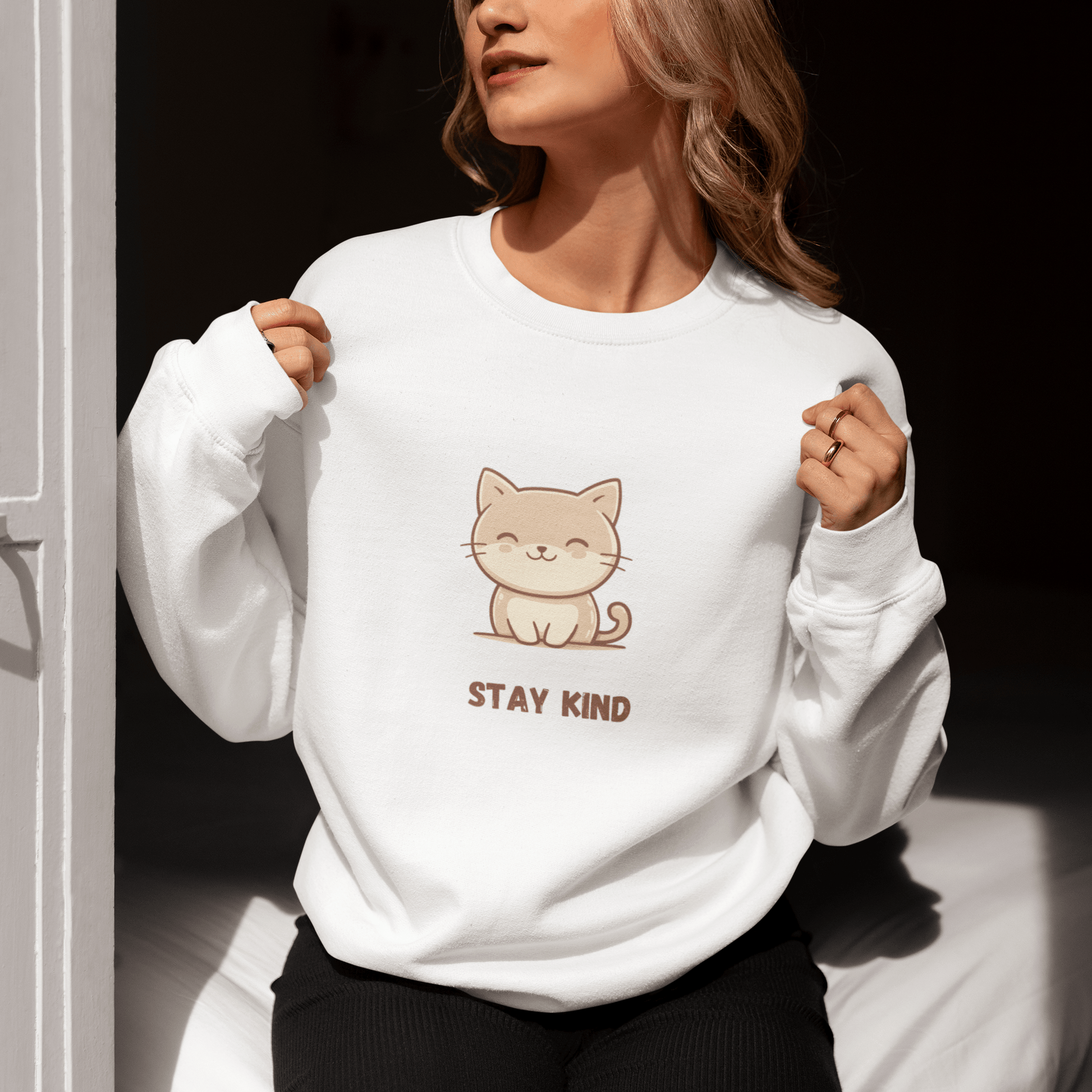 Woman sitting by a sunlit door, wearing a white sweatshirt with a cute cat illustration and the phrase “Stay Kind,” holding the collar with both hands and enjoying the sunlight.