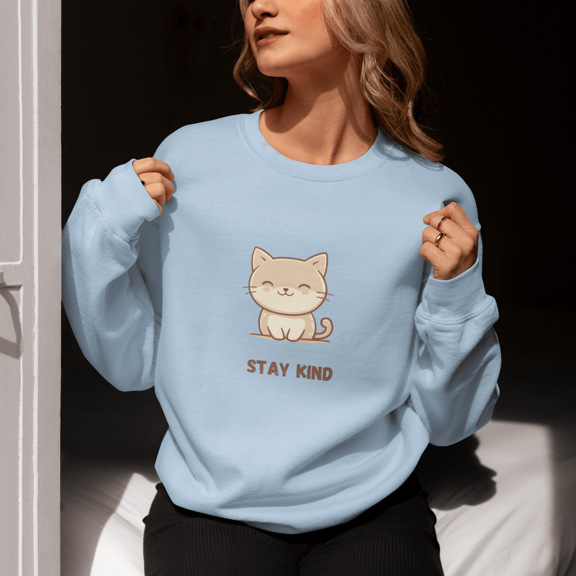 Woman sitting by a sunlit door, wearing a light blue sweatshirt with a cute cat illustration and the phrase “Stay Kind,” holding the collar with both hands and enjoying the sunlight.
