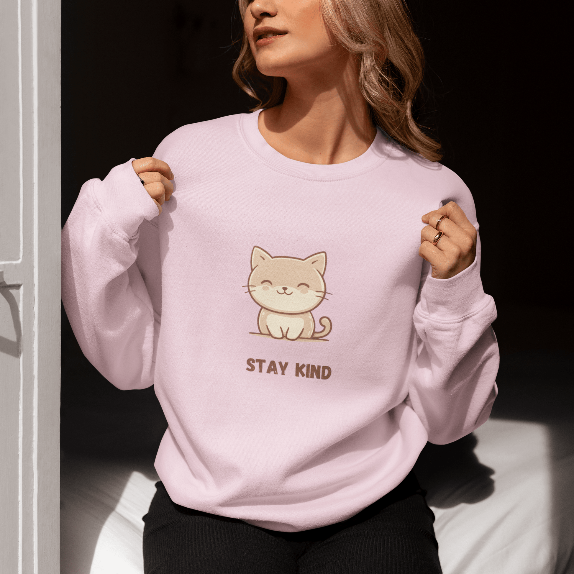 Woman sitting by a sunlit door, wearing a soft pink sweatshirt with a cute cat illustration and the phrase “Stay Kind,” holding the collar with both hands and enjoying the sunlight.
