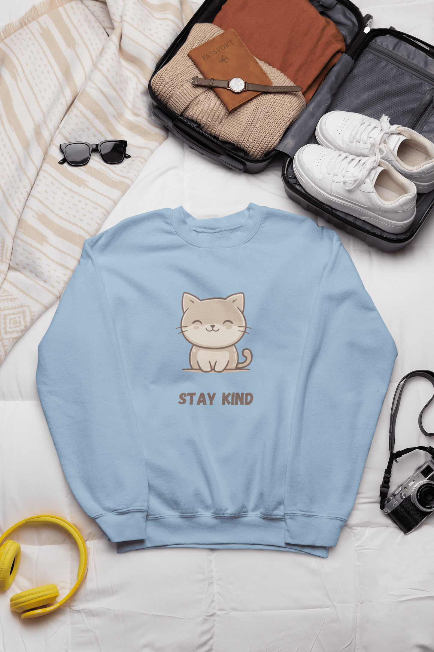 Light blue sweatshirt with a cute cat and the words “Stay Kind” placed on a bed, next to a travel suitcase, sneakers, folded clothes, sunglasses, and travel accessories.
