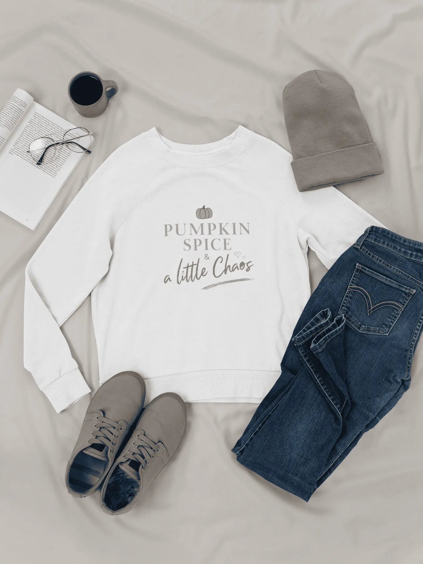 White sweatshirt with 'Pumpkin Spice & a Little Chaos' text, paired with jeans, shoes, and a beanie on a light surface.