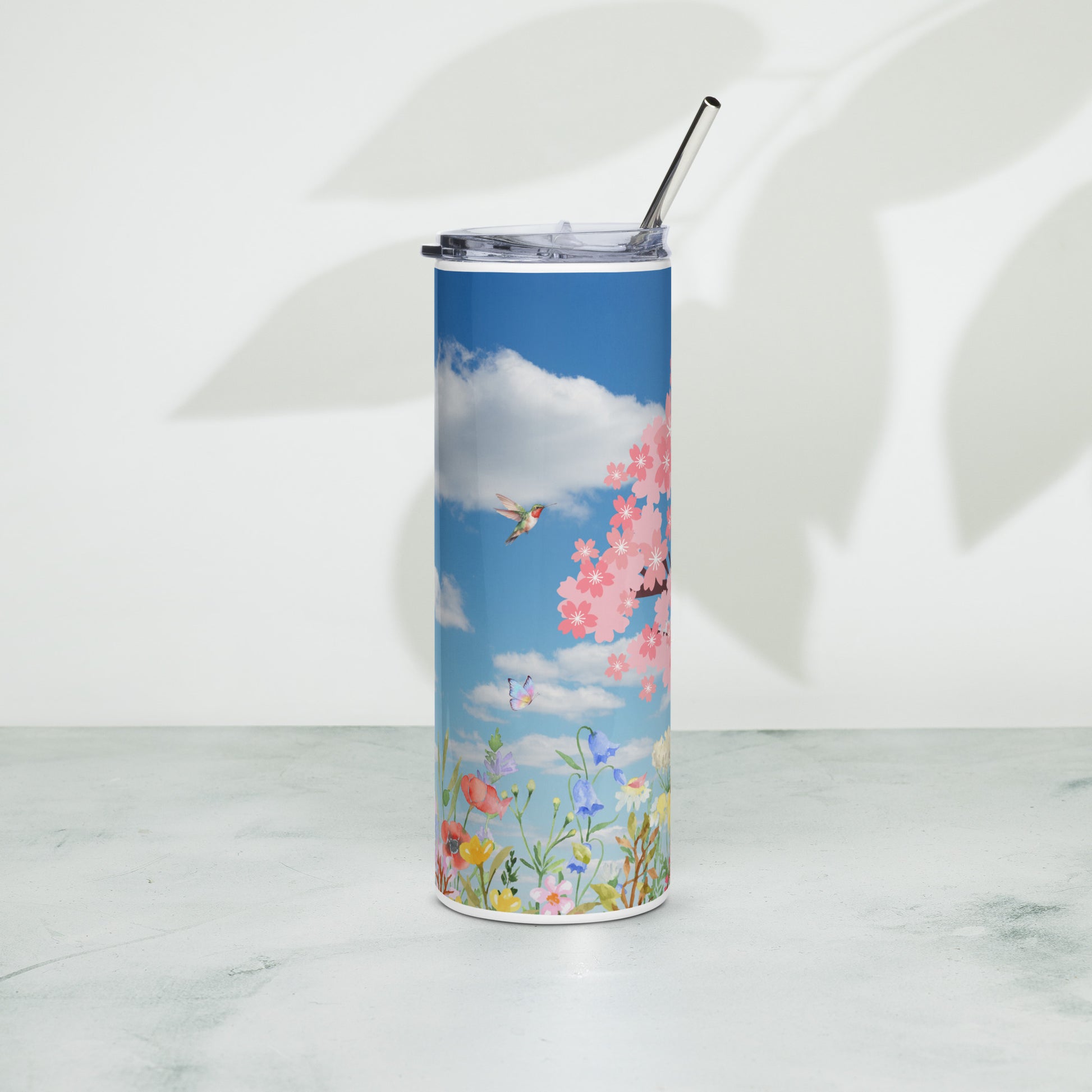 Stainless steel tumbler with a metal straw, featuring a bright blue sky with clouds, cherry blossoms, wildflowers, and butterflies, standing upright on a light surface with soft leaf shadows in the background.