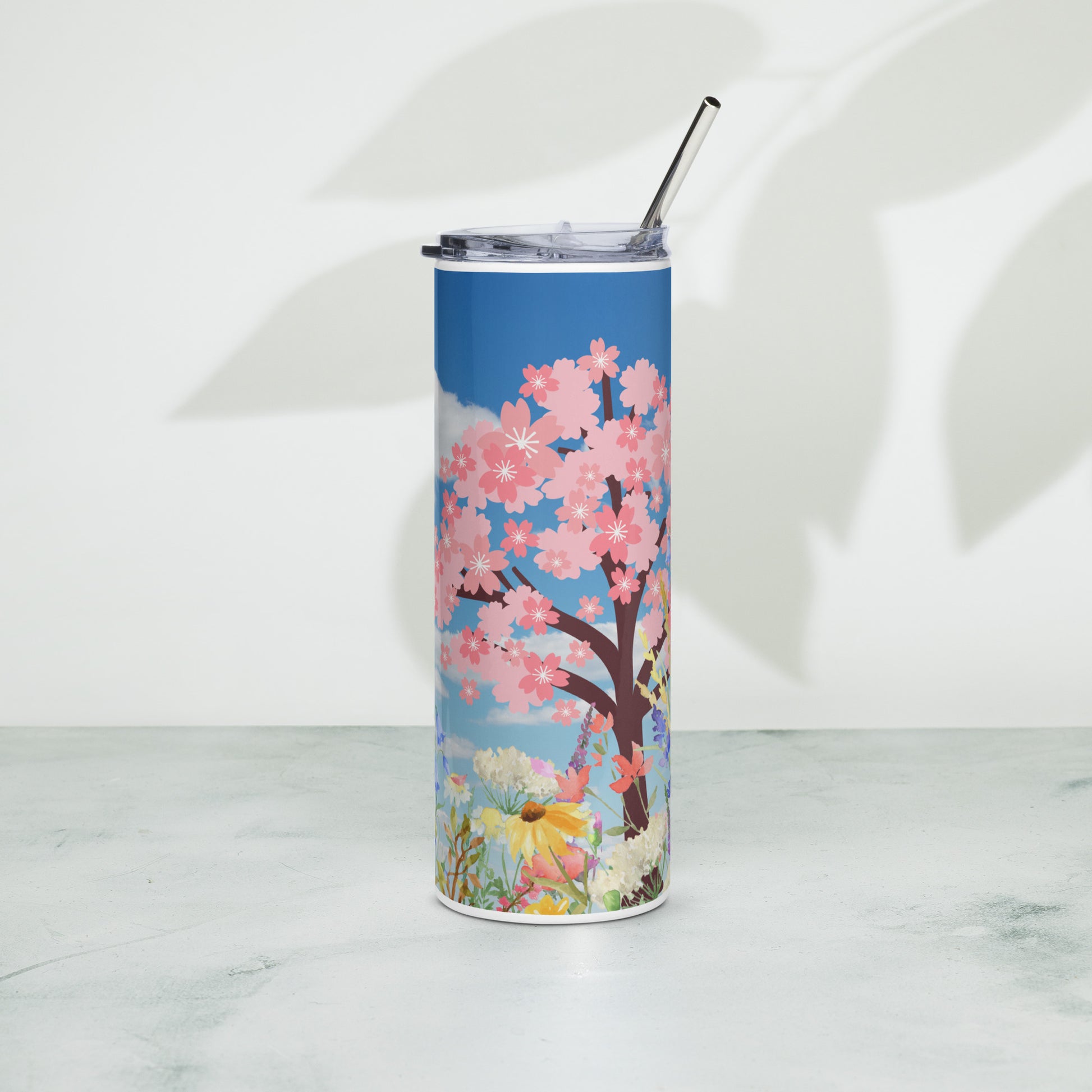 Stainless steel tumbler with metal straw, featuring a vivid cherry blossom tree and pastel floral design on a blue sky background, placed upright on a light surface.
