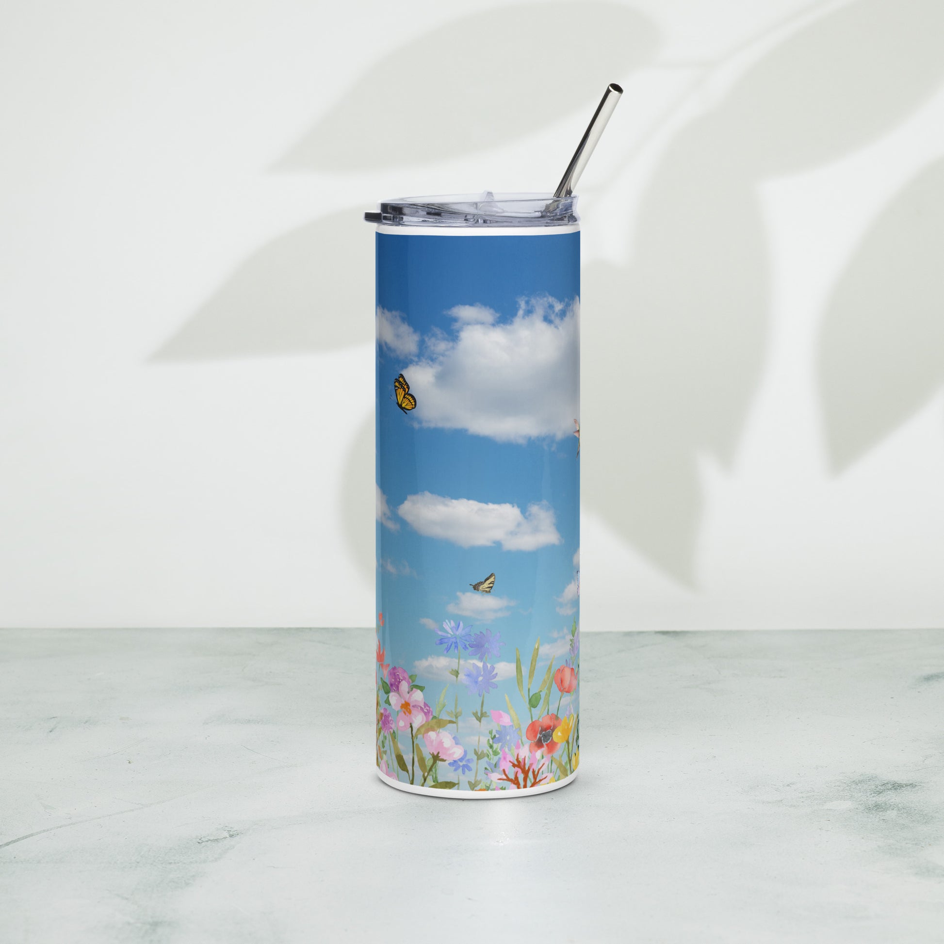 Stainless steel tumbler with a metal straw, decorated with blue sky, fluffy white clouds, butterflies, and colorful spring flowers, standing on a light surface with soft leaf shadows in the background.
