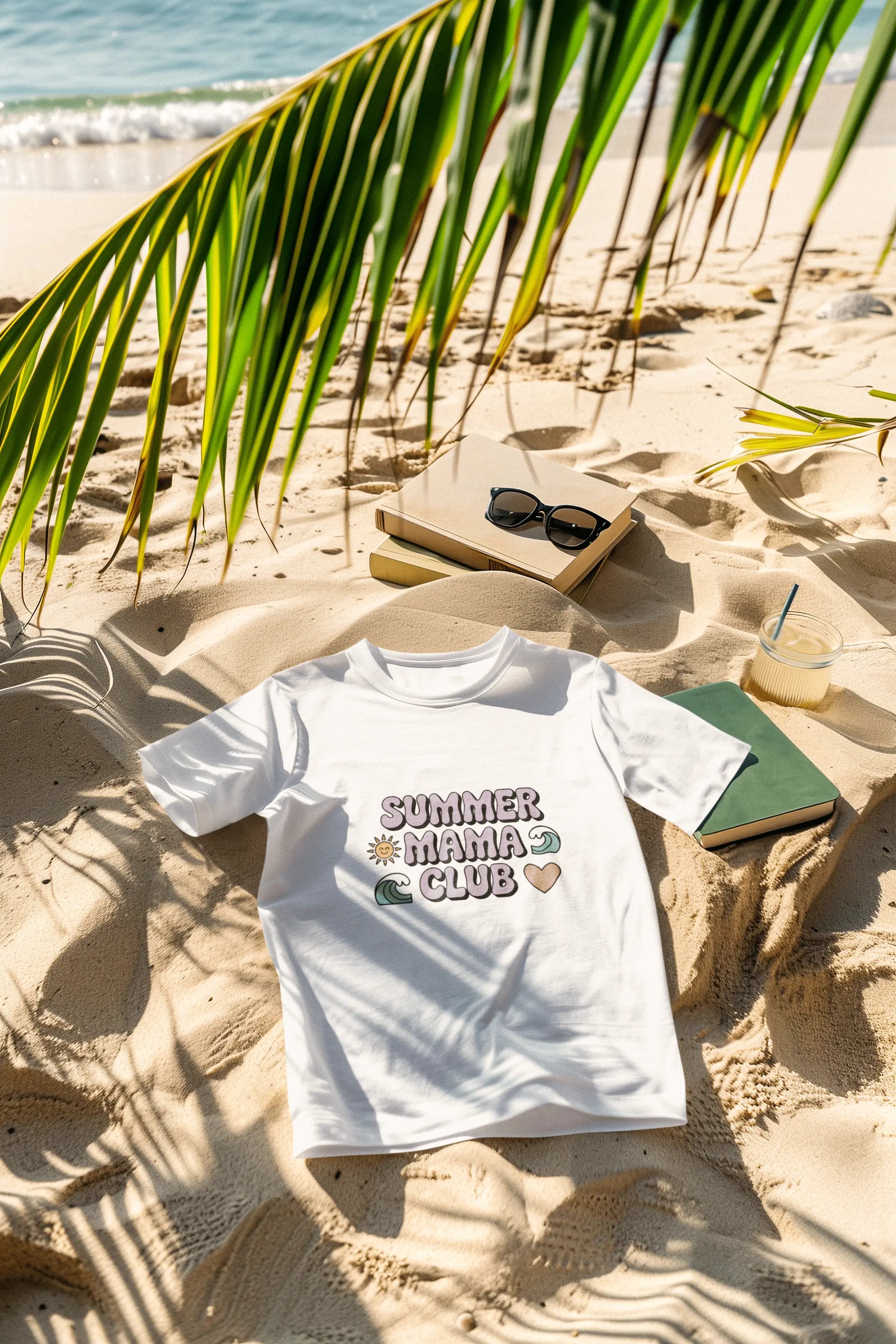 White Summer Mama Club t-shirt on sand with books, sunglasses, and iced coffee – beach flatlay mockup for Happily Styled summer collection