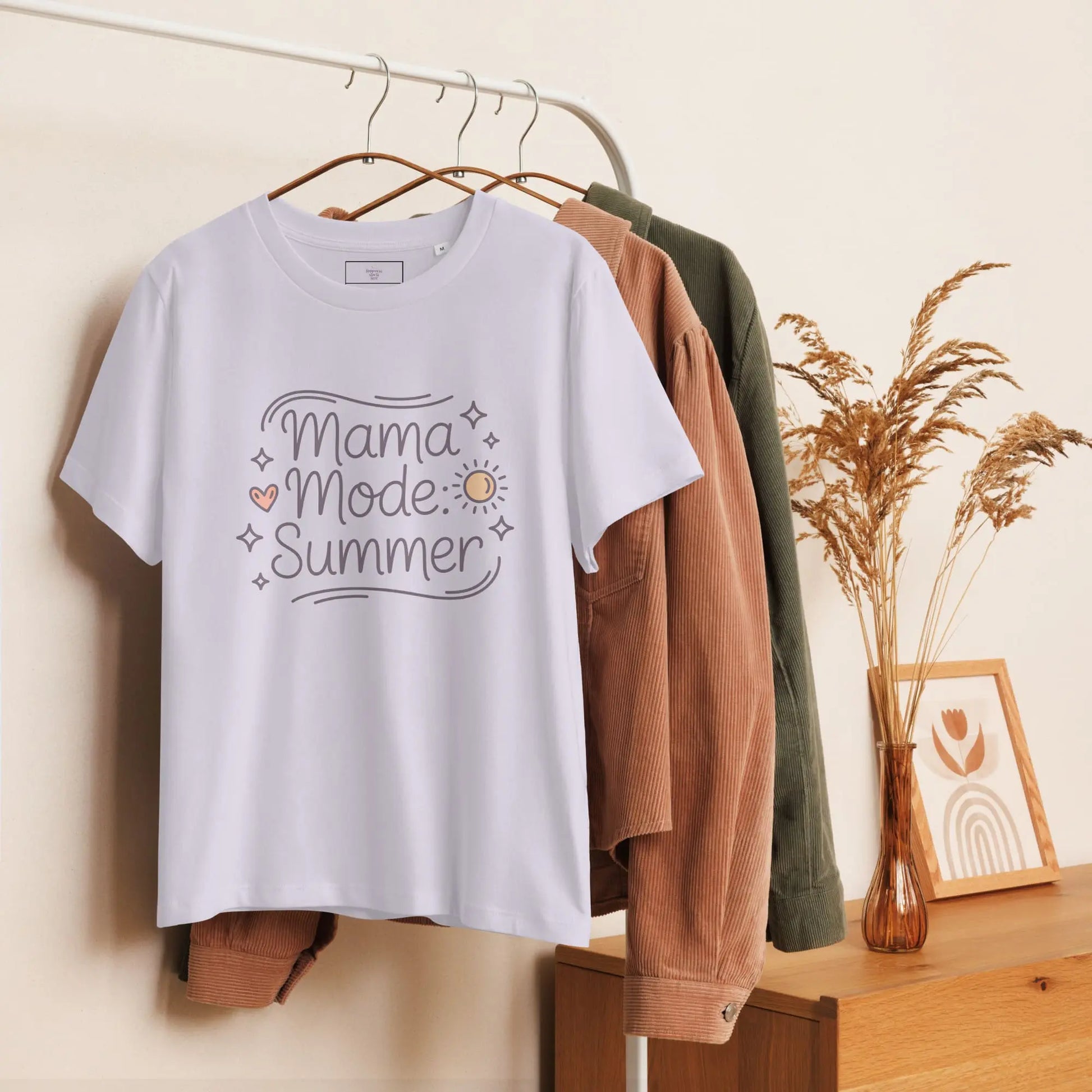 Hanging lavander t-shirt with 'Mama Mode: Summer' design displayed on a wooden clothing rack with neutral decor.