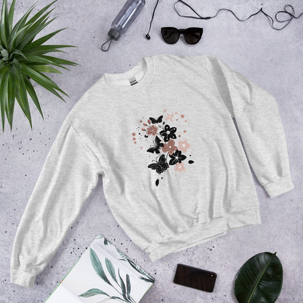 Flat lay of a ash gray sweatshirt with a black and pink floral butterfly design, styled with sunglasses, a water bottle, and leaves on a light stone background.