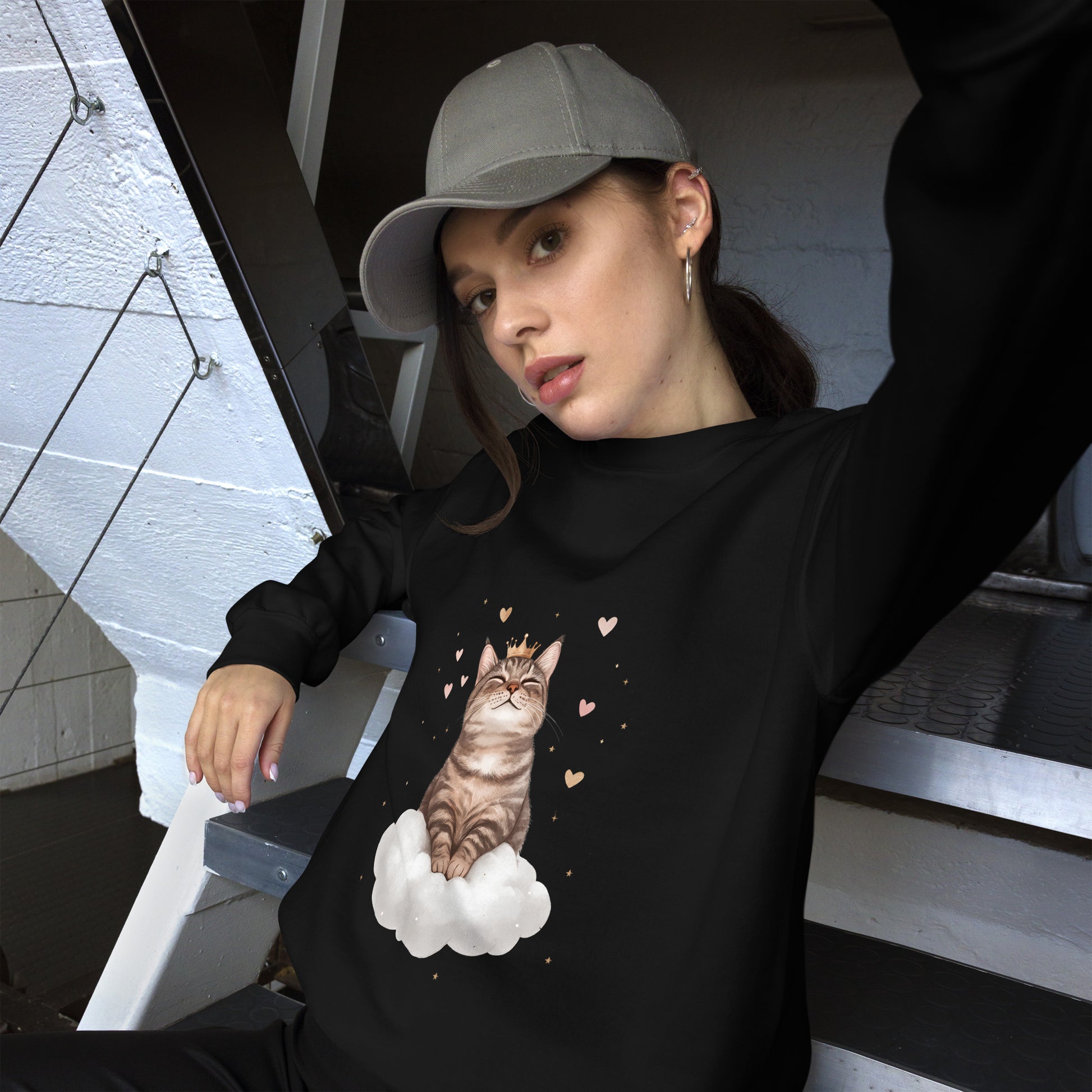 Woman wearing a black sweatshirt with a realistic cat sitting on a white cloud, surrounded by floating hearts. She is sitting casually on stairs, wearing a gray cap and looking relaxed