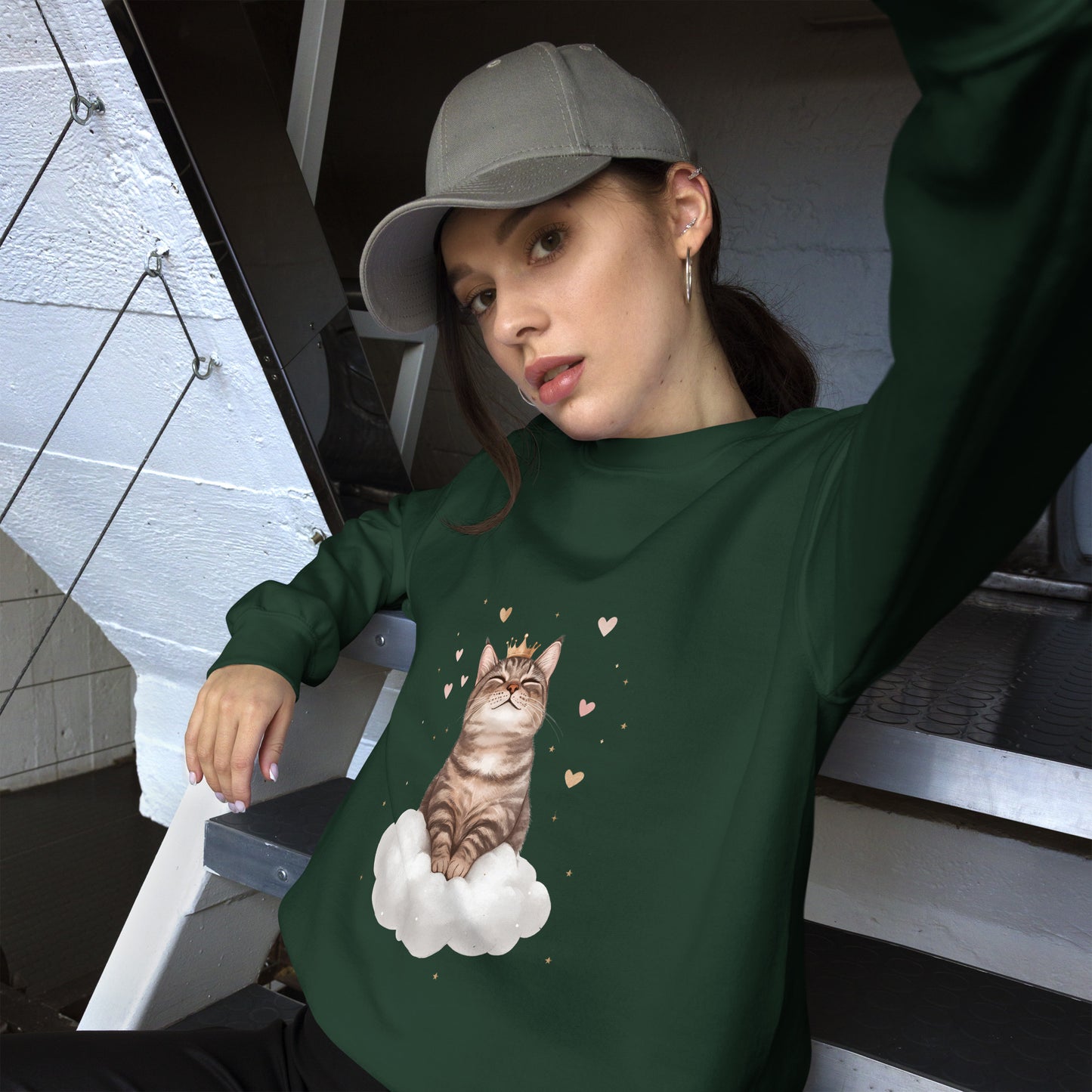 Woman wearing a forest green sweatshirt with a realistic cat sitting on a white cloud, surrounded by floating hearts. She is sitting casually on stairs, wearing a gray cap and looking relaxed
