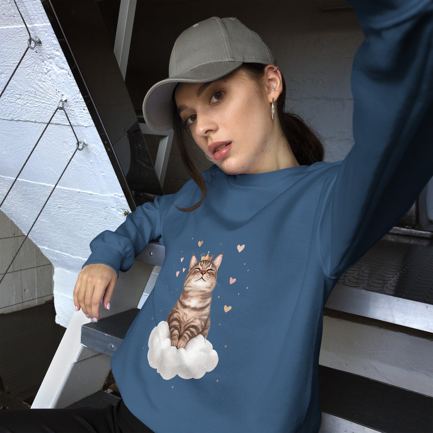 Woman wearing a indigo blue sweatshirt with a realistic cat sitting on a white cloud, surrounded by floating hearts. She is sitting casually on stairs, wearing a gray cap and looking relaxed