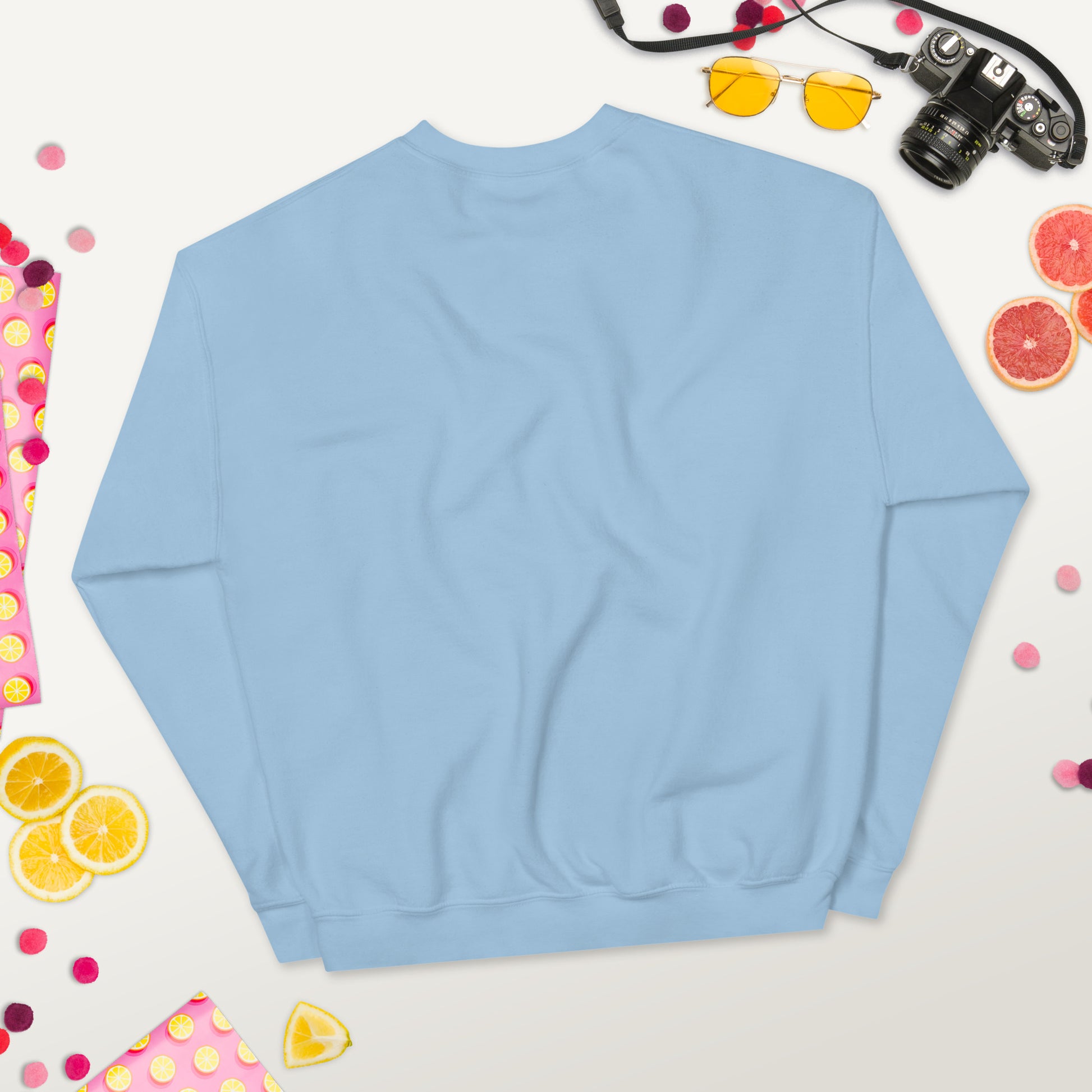 Back view of a light blue unisex crew neck sweatshirt laid flat on a colorful background with fruits, sunglasses, and a camera.