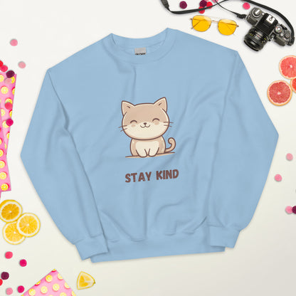 Light blue sweatshirt with a cute cat and the phrase “Stay Kind,” laid flat on a white surface, surrounded by colorful fruits, sunglasses, a camera, and playful decorative elements.