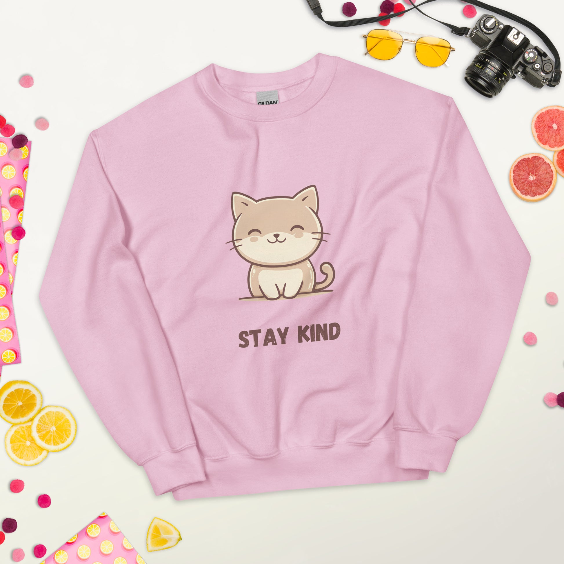 Soft pink sweatshirt with a cute cat and the phrase “Stay Kind,” laid flat on a white surface, surrounded by colorful fruits, sunglasses, a camera, and playful decorative elements.