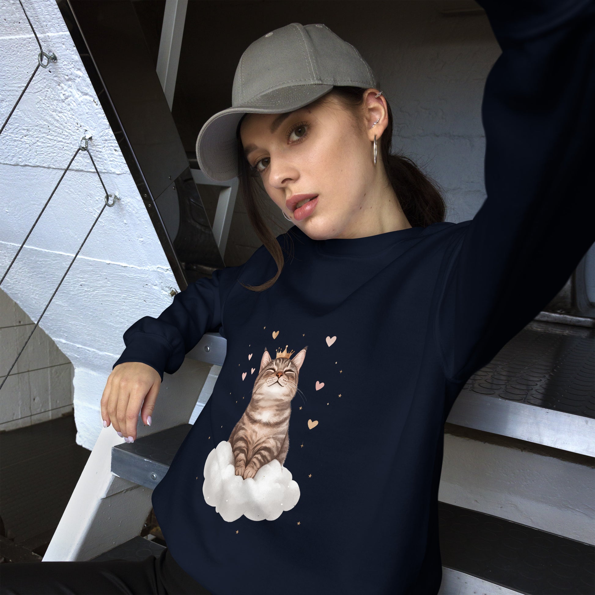 Woman wearing a navy blue sweatshirt with a realistic cat sitting on a white cloud, surrounded by floating hearts. She is sitting casually on stairs, wearing a gray cap and looking relaxed