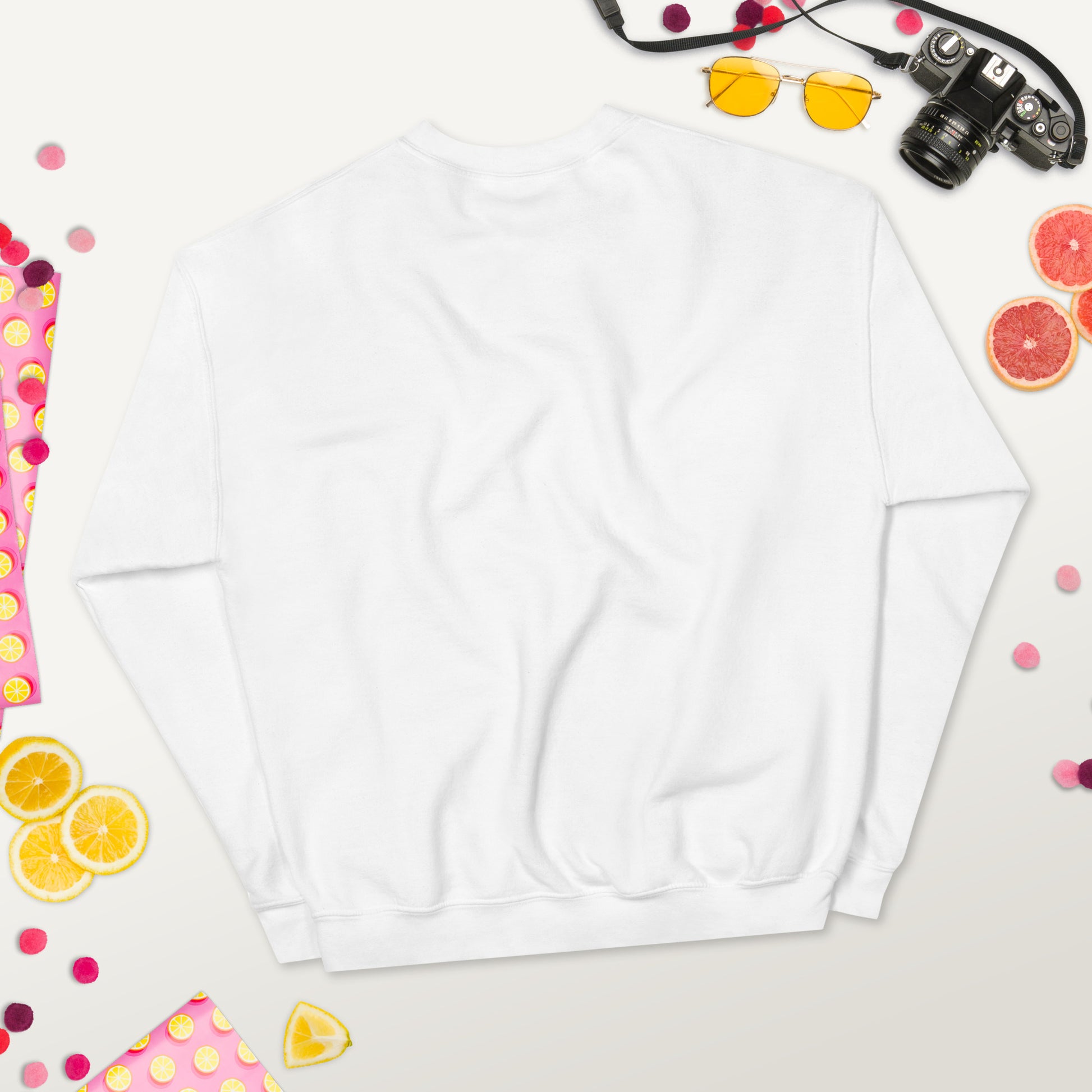 Back view of a white unisex crew neck sweatshirt laid flat on a colorful background with fruits, sunglasses, and a camera.