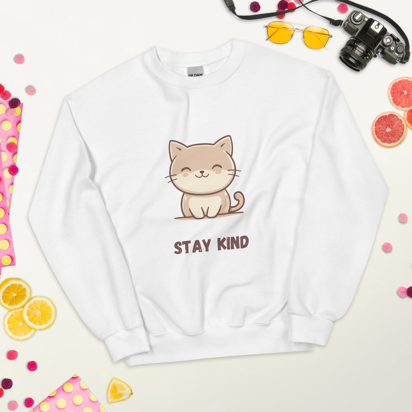 White sweatshirt with a cute cat and the phrase “Stay Kind,” laid flat on a white surface, surrounded by colorful fruits, sunglasses, a camera, and playful decorative elements.