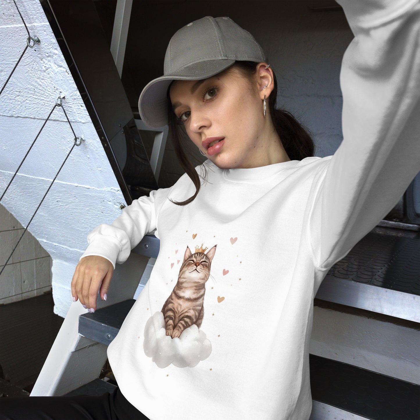 Woman wearing a white sweatshirt with a realistic cat sitting on a white cloud, surrounded by floating hearts. She is sitting casually on stairs, wearing a gray cap and looking relaxed