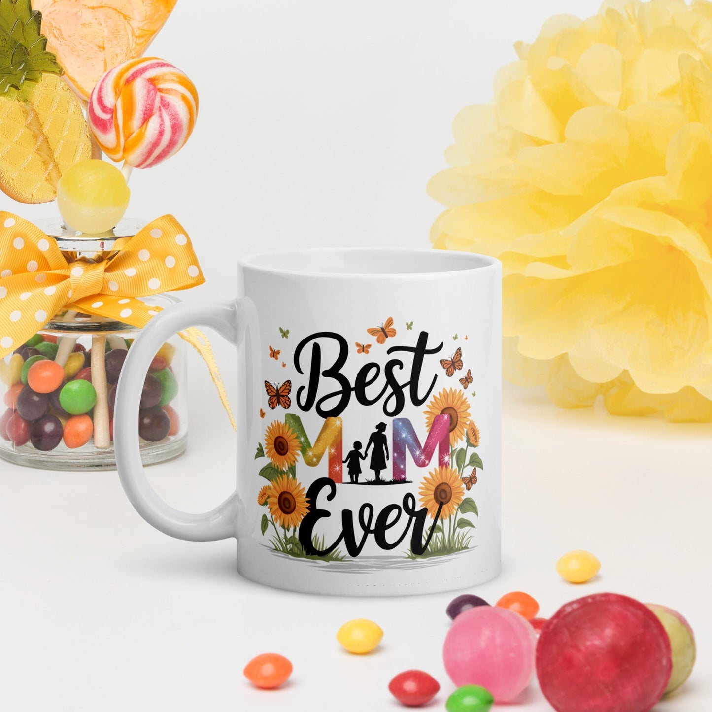 White ceramic mug with floral “Best Mom Ever” design, surrounded by candies and bright yellow decor on a white background.