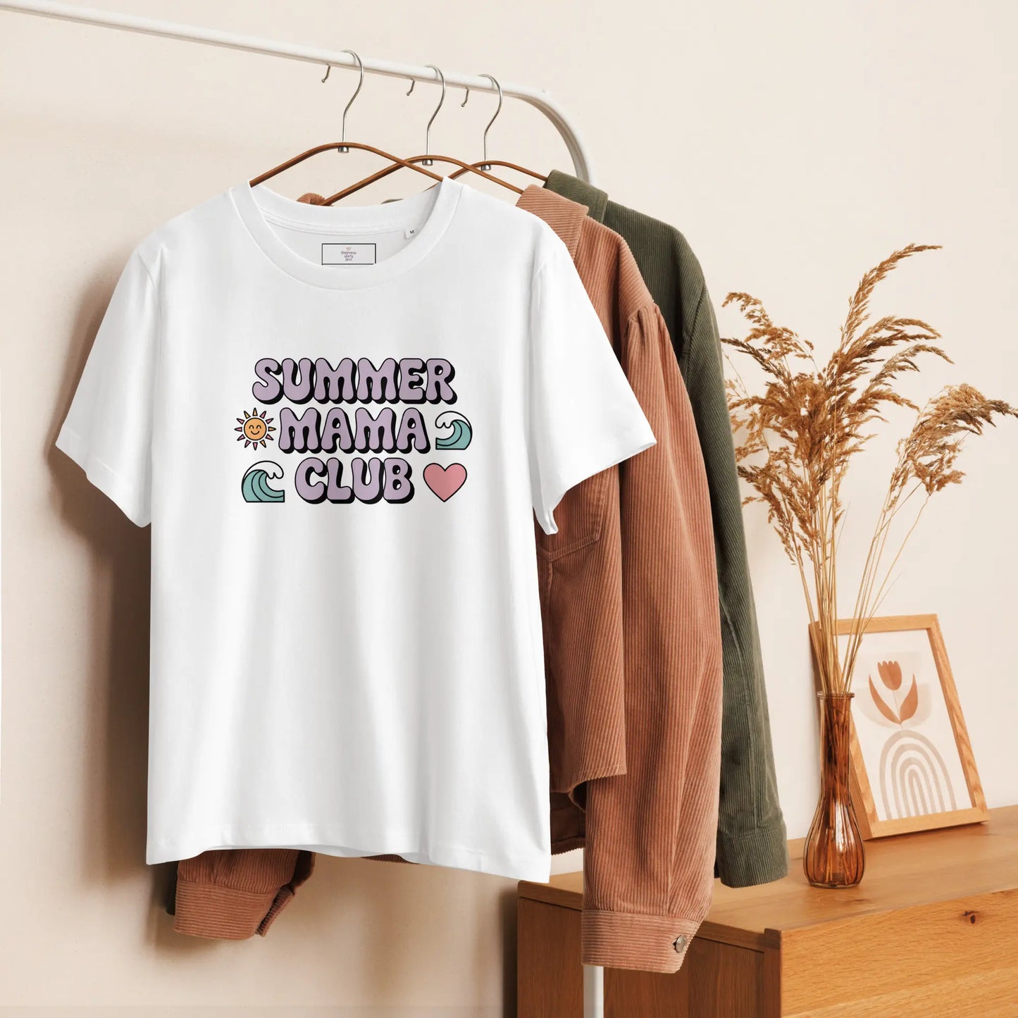 White t-shirt with pastel "Summer Mama Club" design hanging on a white rack with cozy boho decor in the background.